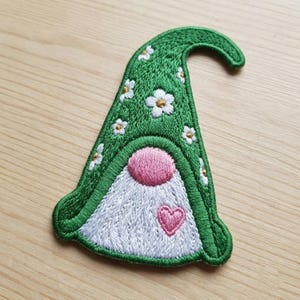 May include: An embroidered patch shaped like a gnome hat in a vibrant green color. The hat features a pattern of small white flowers with yellow centers. Below the hat is a textured white beard, a round pink nose, and a small pink heart.