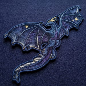 May include: An embroidered patch featuring a dragon in shades of blue and purple with gold-colored thread accents. The dragon is depicted with wings spread wide, showcasing intricate stitching details and small star motifs on the wing surfaces.