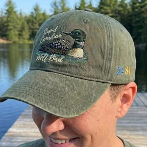 May include: Olive green baseball cap with a curved brim. The cap features an embroidered design of a loon bird and the text "Stupid Canadian Wolf Bird". The number "2481" is embroidered on the side.