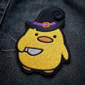 May include: An embroidered patch featuring a round yellow chick wearing a black witch hat with a purple band and an orange buckle. The chick has a small orange beak and is holding a silver-colored knife with a black handle in its wing.