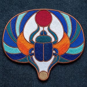 May include: An embroidered patch featuring a central blue scarab beetle with outstretched wings in shades of orange, teal, and blue. A red circular sun sits above the beetle, and a small tan circle is at the bottom, all outlined in copper thread.