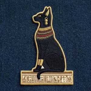 May include: An embroidered patch featuring a black cat sitting in profile on a rectangular base. The cat has gold-colored ear, neck, and leg accents. The base is tan with dark brown hieroglyphic symbols. The patch is set against a dark blue denim fabric.