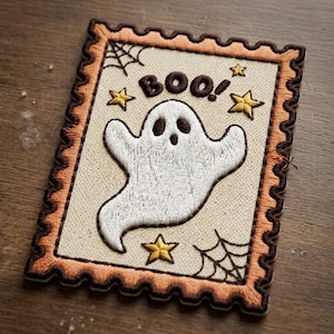 May include: A rectangular embroidered patch shaped like a postage stamp with a cream-colored center. It features a white ghost, the text "BOO!" in dark brown, three yellow stars, and two black spiderwebs, all bordered by a scalloped orange frame.