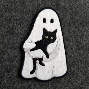 May include: An embroidered patch featuring a white ghost figure with a black outline. The ghost has two small square eyes and is holding a black cat with bright yellow-green eyes. The cat is positioned horizontally across the center of the ghost.
