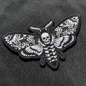 May include: An embroidered patch featuring a moth with a skull pattern on its thorax. The design is rendered in black and white thread with intricate wing details, a segmented abdomen, and feathery antennae, all outlined with a thick black border.