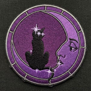 May include: A circular embroidered patch featuring a black cat silhouette sitting on a purple crescent moon with a human-like face. The design includes small white star accents and a segmented border, all rendered in purple, black, and white thread.