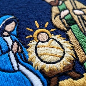 May include: A detailed embroidery on a dark blue fabric background depicting a nativity scene. The design features a central infant in a yellow manger with a radiating sunburst, flanked by a figure in a blue robe and a figure in a tan robe.