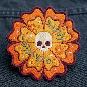 May include: An embroidered patch featuring a central white skull surrounded by yellow and orange flower petals. The design includes small green leafy branches, tiny orange blossoms, and white star accents, all outlined with a dark purple border.