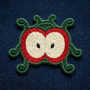 May include: An embroidered patch featuring a stylized green creature with four curled limbs. The center displays two large, circular shapes with red borders, cream-colored interiors, and small black dots, resembling sliced apples or eyes.