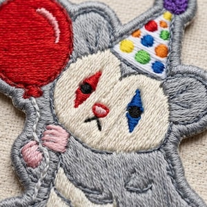 May include: An embroidered patch featuring a gray character wearing a party hat with colorful polka dots. The character has a red diamond shape over one eye, a blue diamond over the other, and a small pink nose. It holds a large red balloon.