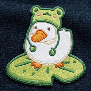 May include: An embroidered patch featuring a white duck with an orange beak wearing a green frog-shaped hat with pink cheeks. The duck is standing on a large, light green lily pad with dark green veins, all outlined in a thick green border.