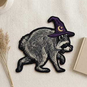 May include: An embroidered patch featuring a gray and black raccoon walking while wearing a purple pointed hat. The hat is decorated with a gold crescent moon and a small star. The patch has a distinct black border and detailed stitching.