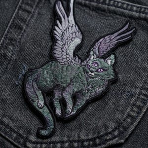 May include: An embroidered patch featuring a winged cat with three eyes. The cat is depicted in shades of dark green and purple, with detailed stitching creating a textured appearance. The wings are spread wide, and the cat has a long tail.
