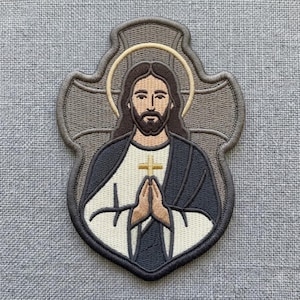 May include: An embroidered patch featuring a figure with long brown hair and a beard, wearing a white robe and a dark gray cloak. The figure has hands pressed together in prayer, holding a small gold cross, set against a gray cross-shaped background.