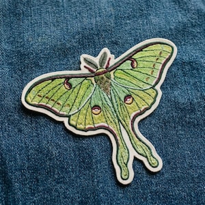 May include: An embroidered patch shaped like a Luna moth with light green wings, dark green veins, and maroon accents. The moth features feathery antennae, a fuzzy body, and long, trailing hind wings with circular eye-spot markings in yellow and black.