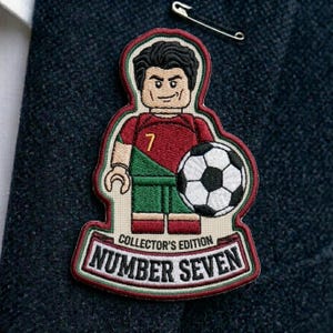 May include: An embroidered patch featuring a stylized minifigure wearing a red and green soccer jersey with the number 7, holding a black and white soccer ball. The patch includes the text COLLECTOR'S EDITION and NUMBER SEVEN in black lettering.