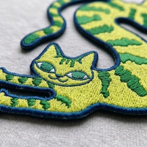May include: An embroidered patch featuring a stylized cat with light green fur and dark green stripes. The cat has a dark blue stitched border, almond-shaped green eyes with white accents, and a curved tail that extends upward and to the right.