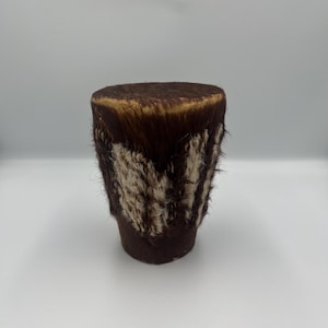 May include: A small, cylindrical drum with a dark brown, fur-covered top and sides. The sides feature a band of cream-colored fur with dark brown accents. The drum is likely handmade and has a rustic appearance.