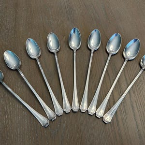 Könnte beinhalten: Neun silberne Löffel auf einer dunklen Holzoberfläche angeordnet. Die Löffel haben lange, schlanke Stiele und ovale Laffen. Die Griffe haben ein dekoratives Muster am Ansatz. Die Löffel sind reflektierend.