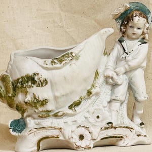 May include: Antique porcelain planter in the shape of a seashell, with a young boy figure in a sailor suit. The planter is white with green and gold accents, and the boy wears a blue hat.