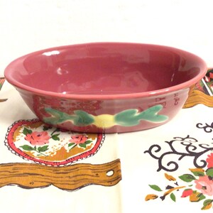 May include: A maroon ceramic bowl with a decorative green and yellow leaf design. The bowl is oval-shaped and sits on a floral patterned tablecloth. The bowl is a vintage kitchenware item.