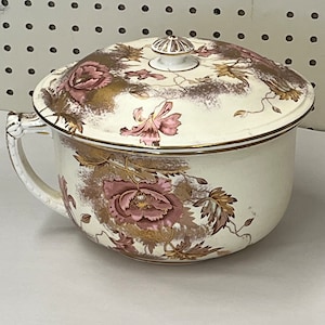 May include: Cream-colored antique chamber pot with a lid. The pot features a floral design in shades of pink and gold, with a gold trim. It has a handle on one side and a decorative knob on the lid.