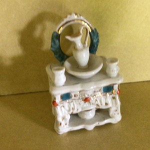 May include: White porcelain miniature vanity set with a gold-trimmed mirror, a pitcher, a basin, and two small jars. The vanity has a blue and gold trim and red accents.