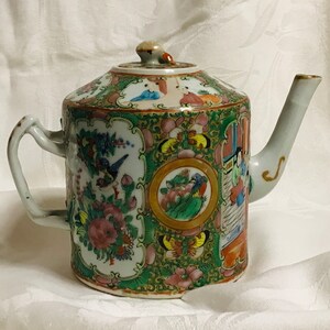 May include: A white porcelain teapot with a green, pink, and gold floral design. The teapot has a round body and a spout on the side. The lid has a knob on top.
