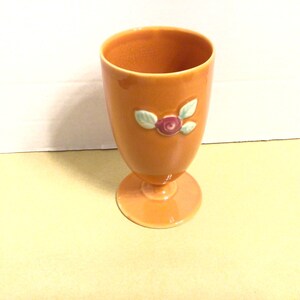 May include: Orange ceramic goblet with a pedestal base. The goblet features a floral design with a red rose and green leaves. The goblet is approximately 6 inches tall and is suitable for decorative use.