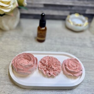 May include: A white oval tray holds three pink floral decorations. The flowers include a peony, a carnation, and a rose. A small brown bottle with a dropper is behind the tray. A white and gold decorative bowl is in the background.