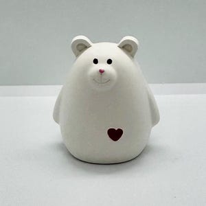 Può includere: Statuetta di orso in ceramica bianca con corpo arrotondato, piccole orecchie e viso sorridente con dettagli rosa. Un cuore rosso è sul petto dell'orso. L'orso è un oggetto decorativo.