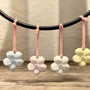 May include: Four flower-shaped ornaments in pastel colours hang from a black curved bar with pink cords. The flowers are light blue, pink, white, and yellow. The background is a light beige colour.