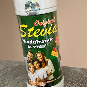 Peut inclure: Un récipient vert et blanc de Stevia Original. L'étiquette présente le mot "Stevia" en écriture rouge et jaune, avec la phrase "Endulzando la vida" en dessous. Un portrait de famille est également sur l'étiquette.