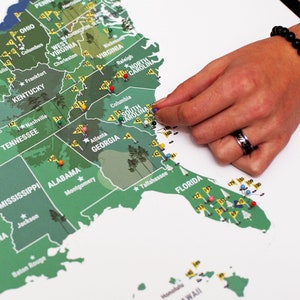 May include: A green and white map of the United States with push pins marking locations. The map is on a white background. The map is a scratch-off map, and the states are outlined in green. The map is a fun way to track your travels.