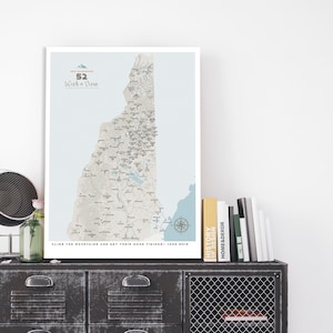 May include: A framed map print of New Hampshire, with the text "52 With a View" and a quote by John Muir. The print is displayed on a dark metal cabinet with drawers and a wire mesh door. Books are stacked on top.