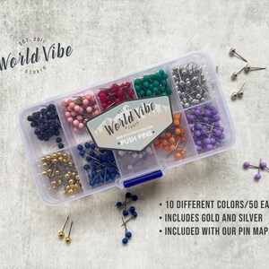 May include: A clear plastic container filled with 10 different colors of push pins, each color has 50 pins. The container has a label that says "World Vibe" and "Push Pins". The container is sitting on a white surface.  The text on the label reads: "World Vibe" and "Push Pins".