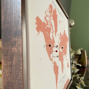 May include: A framed world map with a peach-colored background. The map is mounted on a canvas and has push pins in various colors marking locations.