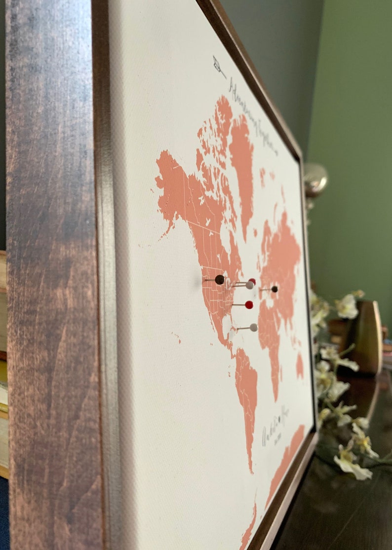 May include: A framed world map with a peach-colored background. The map is mounted on a canvas and has push pins in various colors marking locations.