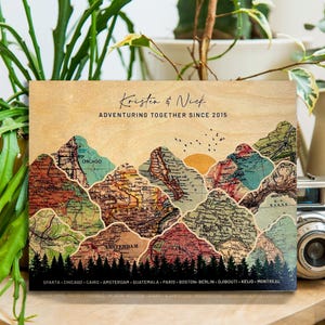 May include: A wooden sign with a vintage map design featuring mountains and a sunrise. The text "Kristen & Nick Adventuring Together Since 2015" is printed at the top. The names of cities are listed at the bottom of the sign.