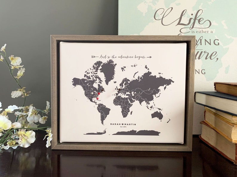 May include: A framed world map with gray countries and a white background. The map is in a wooden frame and has red push pins marking locations. The text "And so the adventure begins" is printed above the map. The text "SARAH & MARTIN Est. 2018" is printed below the map.