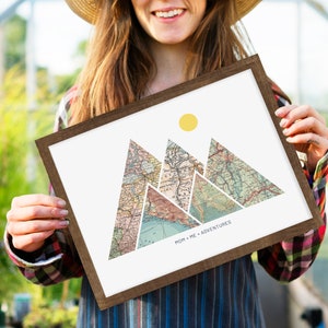 May include: A framed print with a design of three mountains made from vintage maps. The mountains are surrounded by a white background with a yellow sun at the top. The text "MOM + ME + ADVENTURES" is printed at the bottom of the print.