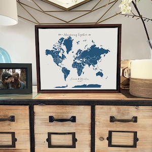 May include: A framed world map with a blue outline of the continents and a white background. The map is titled "Adventuring Together" and has the names "Jared & Martin" and the date "2023" written below the map. The map is sitting on a wooden shelf with a picture frame and a candle.