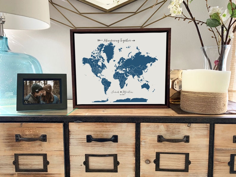 May include: A framed world map with a blue outline of the continents and a white background. The map is titled "Adventuring Together" and has the names "Jared & Martin" and the date "2023" written below the map. The map is sitting on a wooden shelf with a picture frame and a candle.