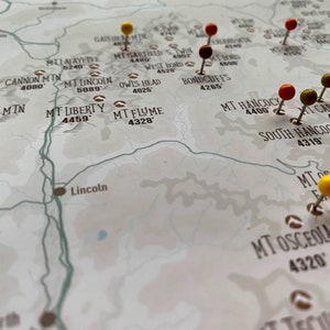 May include: A close-up of a map with push pins marking various locations. The map is printed on white paper with brown text and lines. The push pins are yellow, red, and green.