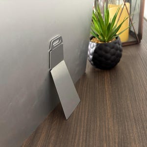 May include: A silver metal picture frame with a built-in easel back. The frame is displayed on a dark wood surface, with a small potted plant and candle in the background. The easel back allows the frame to stand upright.