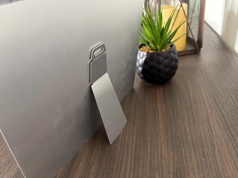 May include: A silver metal picture frame with a built-in easel back. The frame is displayed on a dark wood surface, with a small potted plant and candle in the background. The easel back allows the frame to stand upright.