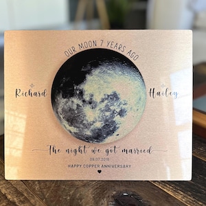 May include: A copper-colored metal sign with a black and white image of the moon. The text on the sign reads "Our moon 7 years ago * Richard Hailey The night we got married 09.07.2018 Happy Copper Anniversary".