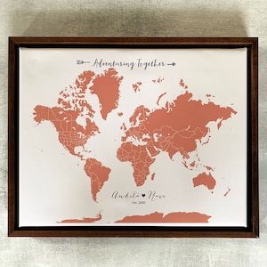 May include: A framed world map with a peach-colored outline of the continents. The map is on a white background with the text "Adventuring Together" and "Ankita & Ravi Est. 2020" printed below the map.