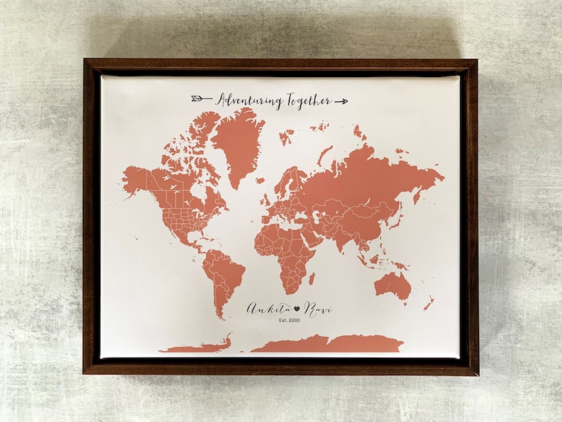 May include: A framed world map with a peach-colored outline of the continents. The map is on a white background with the text "Adventuring Together" and "Ankita & Ravi Est. 2020" printed below the map.