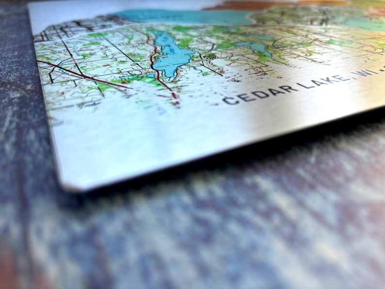 May include: A close-up of a map of Cedar Lake, WI. The map is printed on a white background with blue, green, and brown lines and markings. The text "CEDAR LAKE, WI" is printed in black at the bottom of the map.